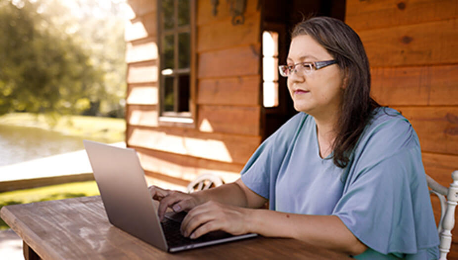 Woman working at a laptop outside on a wooden porch with trees in the background