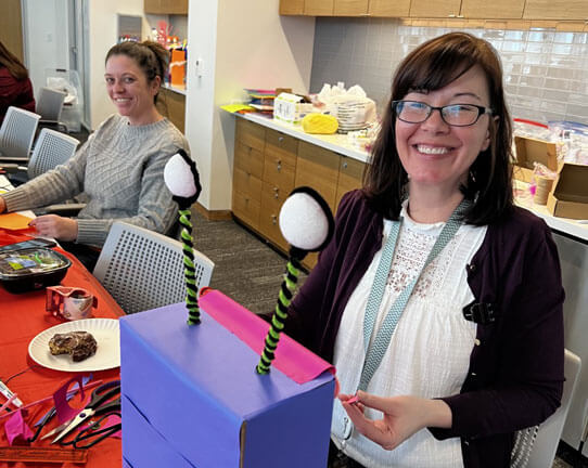 Two women are sitting at a table with colorful craft objects decorating boxes