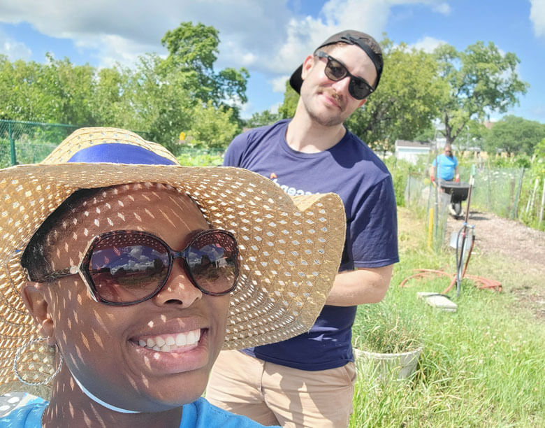 A woman in a sun hat is close to the camera with a man wearing a backwards hat peeking out from behind her. They are outside and it is sunny.