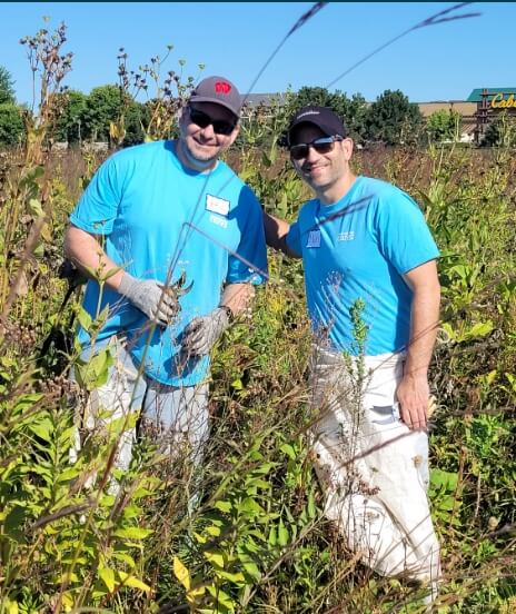 Two men in light blue shirts are stand outside on a sunny day surrounded by tall weeds