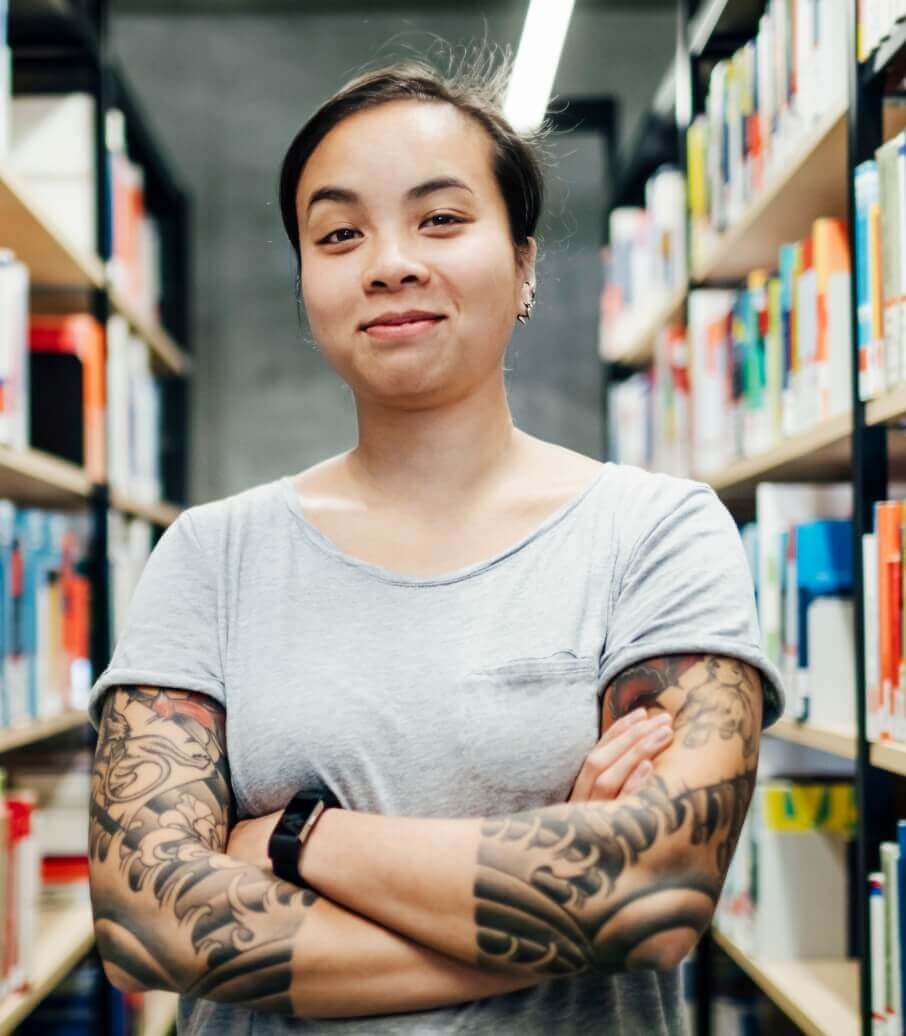 Young woman with tattooed arms standing with arms crossed and smiling in a library