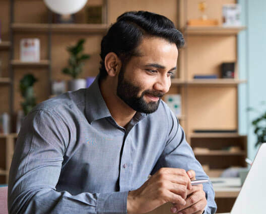 Young man with a beard smiling in front of a laptop in front of neatly arranged shelving