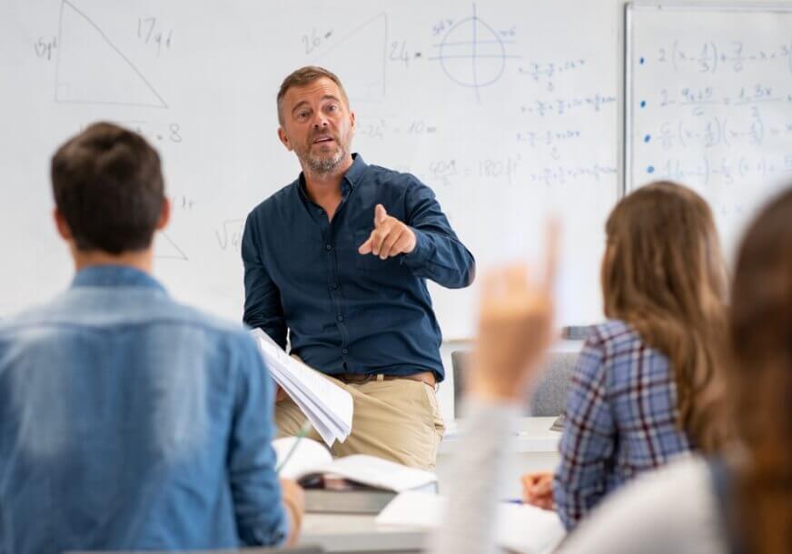 Teacher standing in front of a white board with equations written on it answering a students question