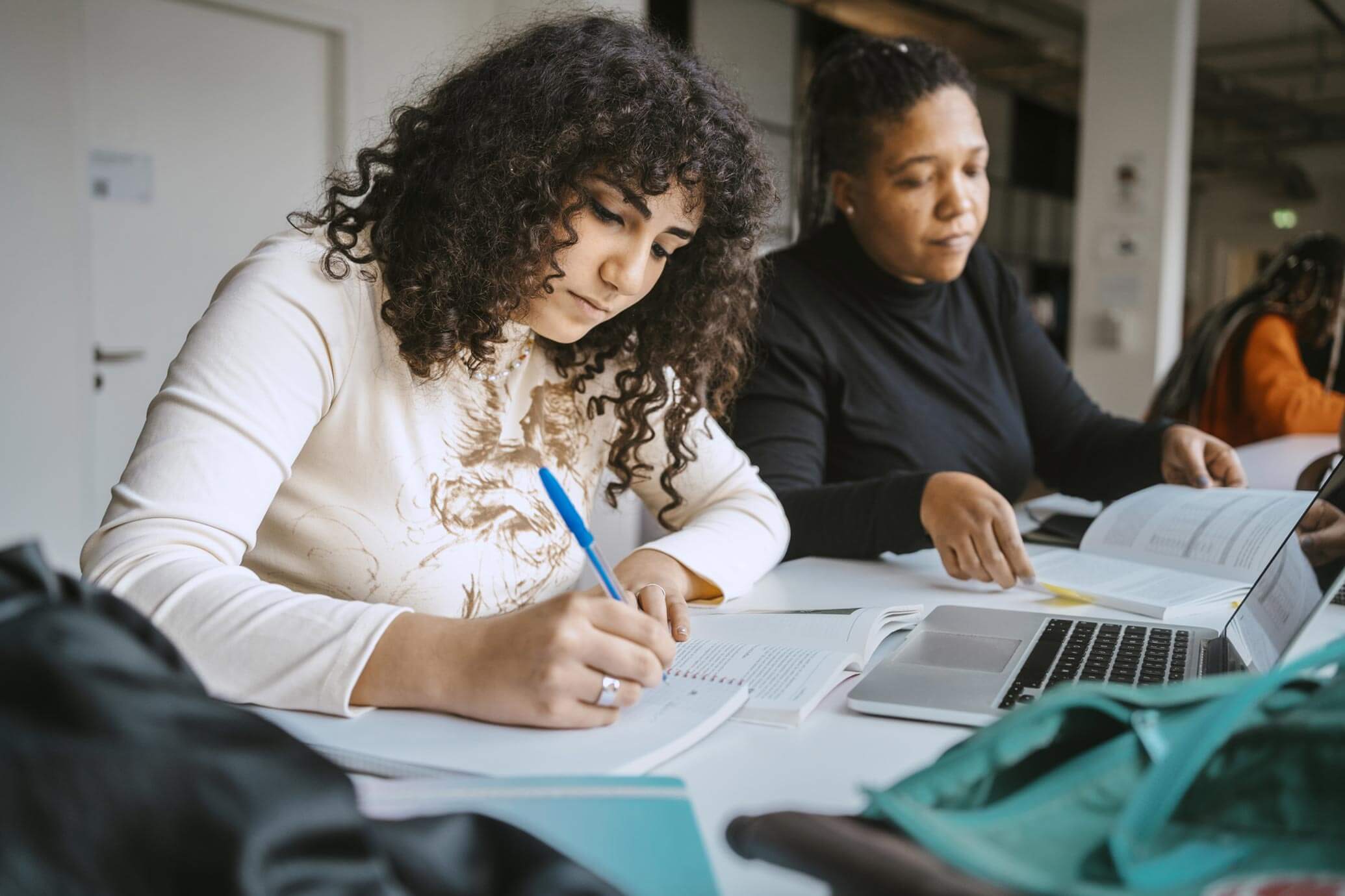 A young woman with dark curly hair is sitting at a table writing with a blue pen. An older woman sits beside her paging though a book