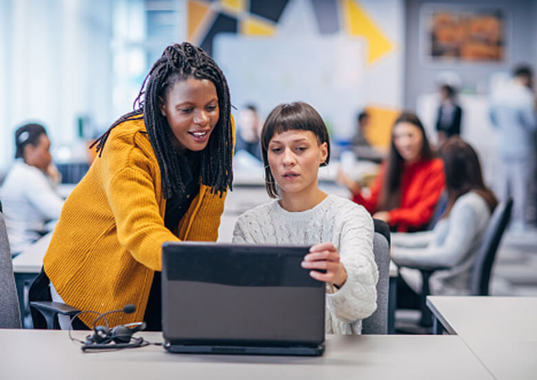 Young woman in classroom with a laptop and a teacher leaning over pointing to the screen